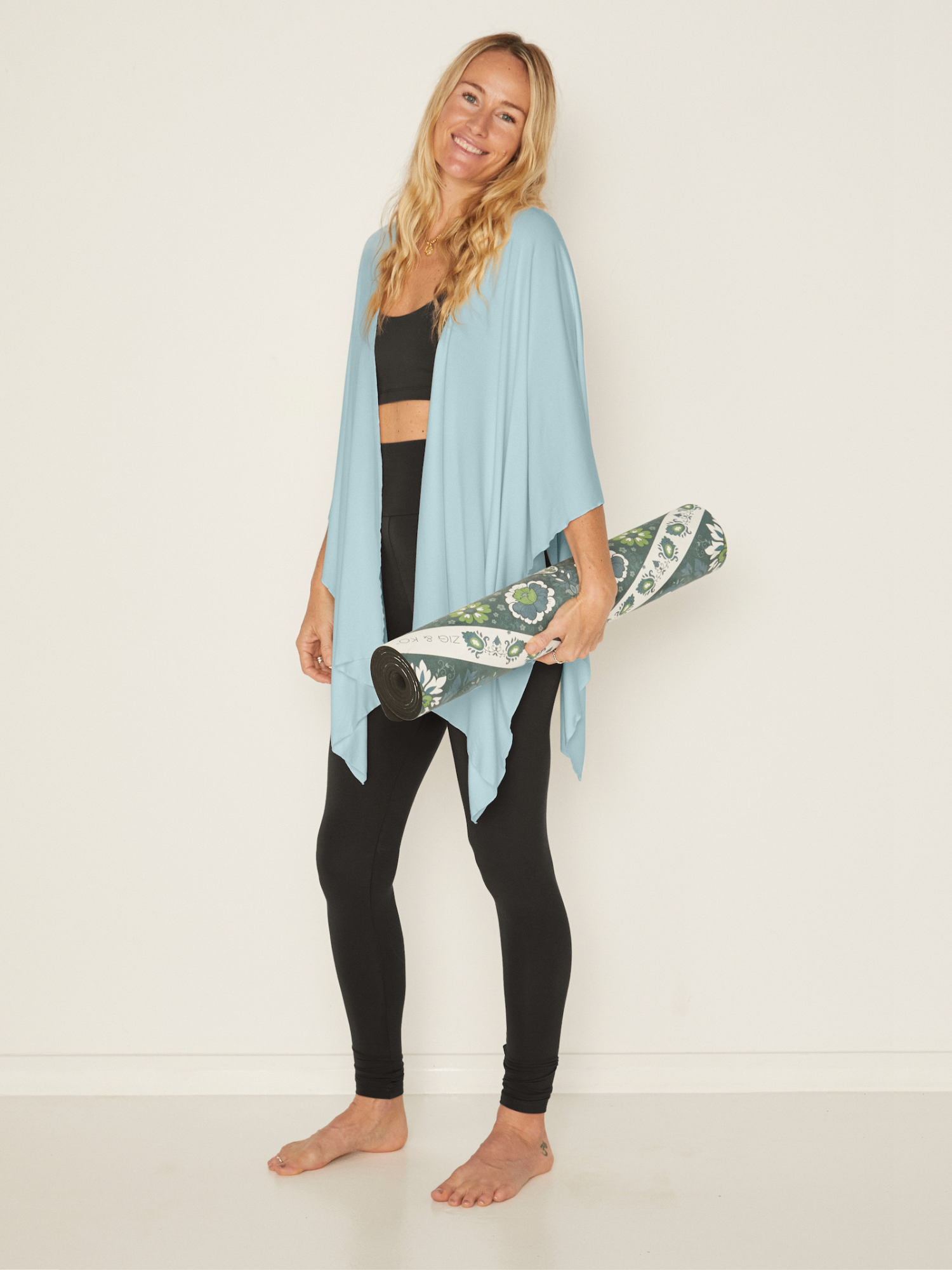 Woman holding a yoga mat with a light blue shawl over her shoulders on a white background