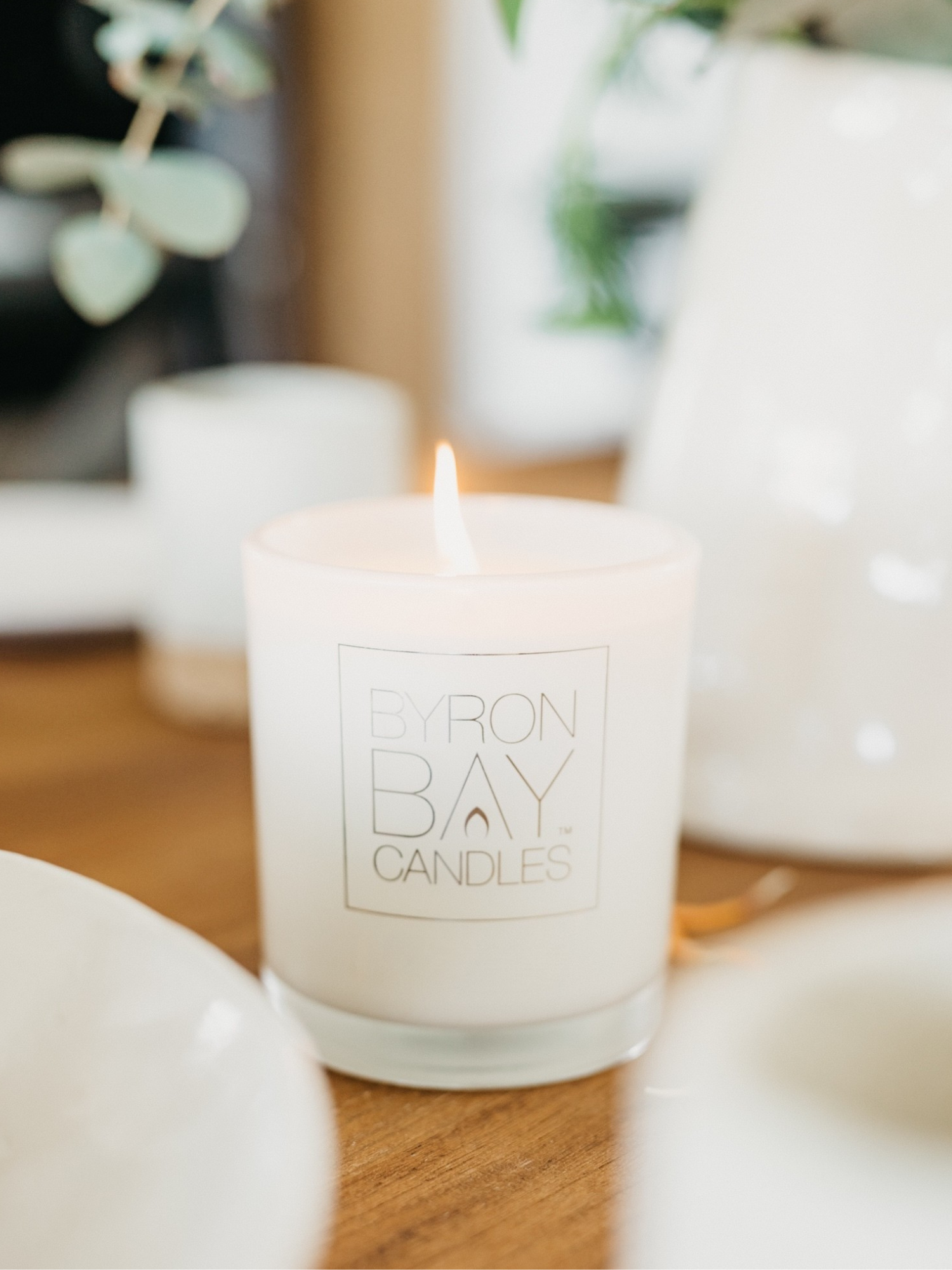 Lit candle with ' Byron Bay Candles' logo on a wooden surface, surrounded by blurred greenery.