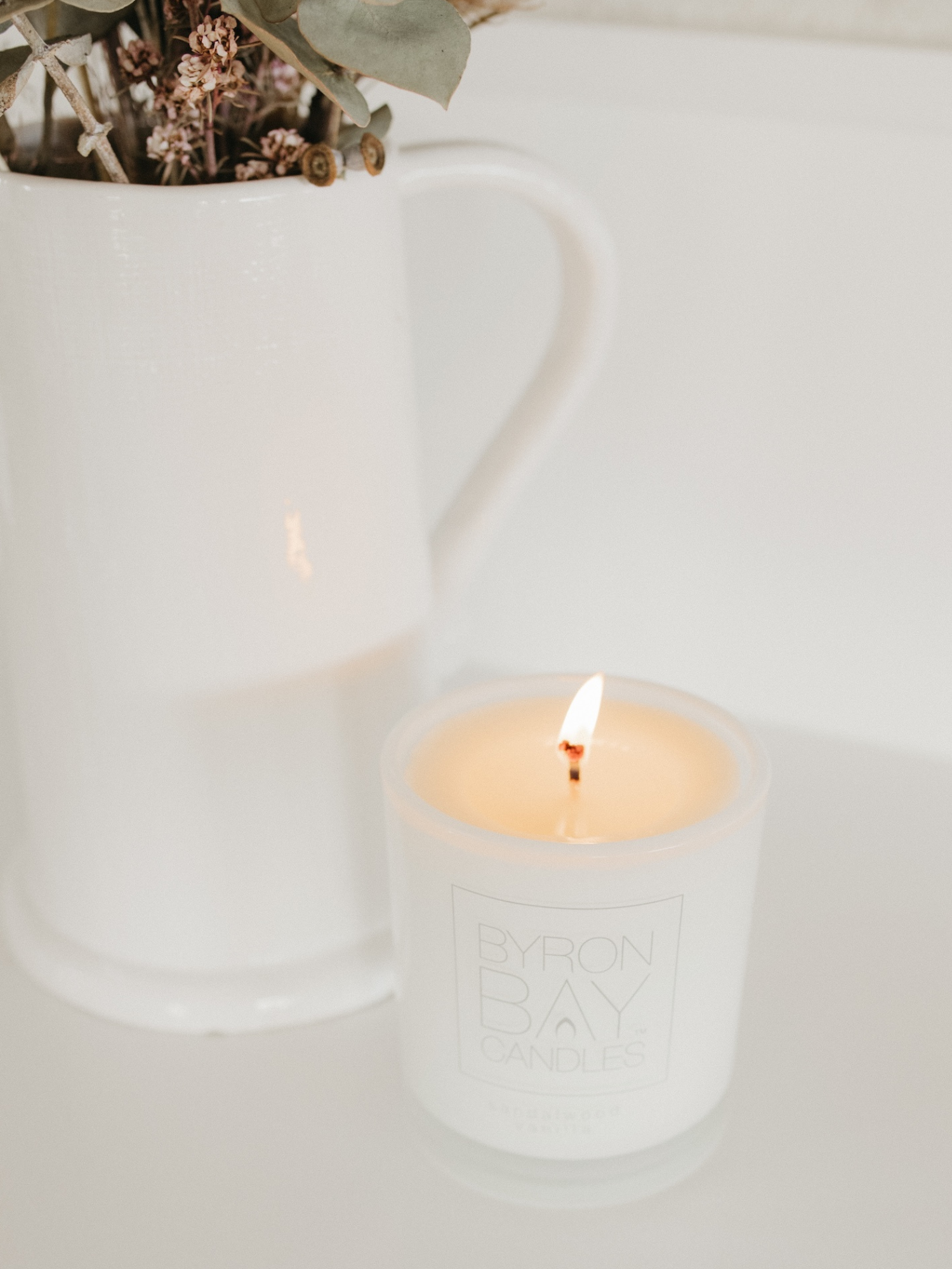 White Byron Bay candle in a glass holder with a vase with dry flowers in the background