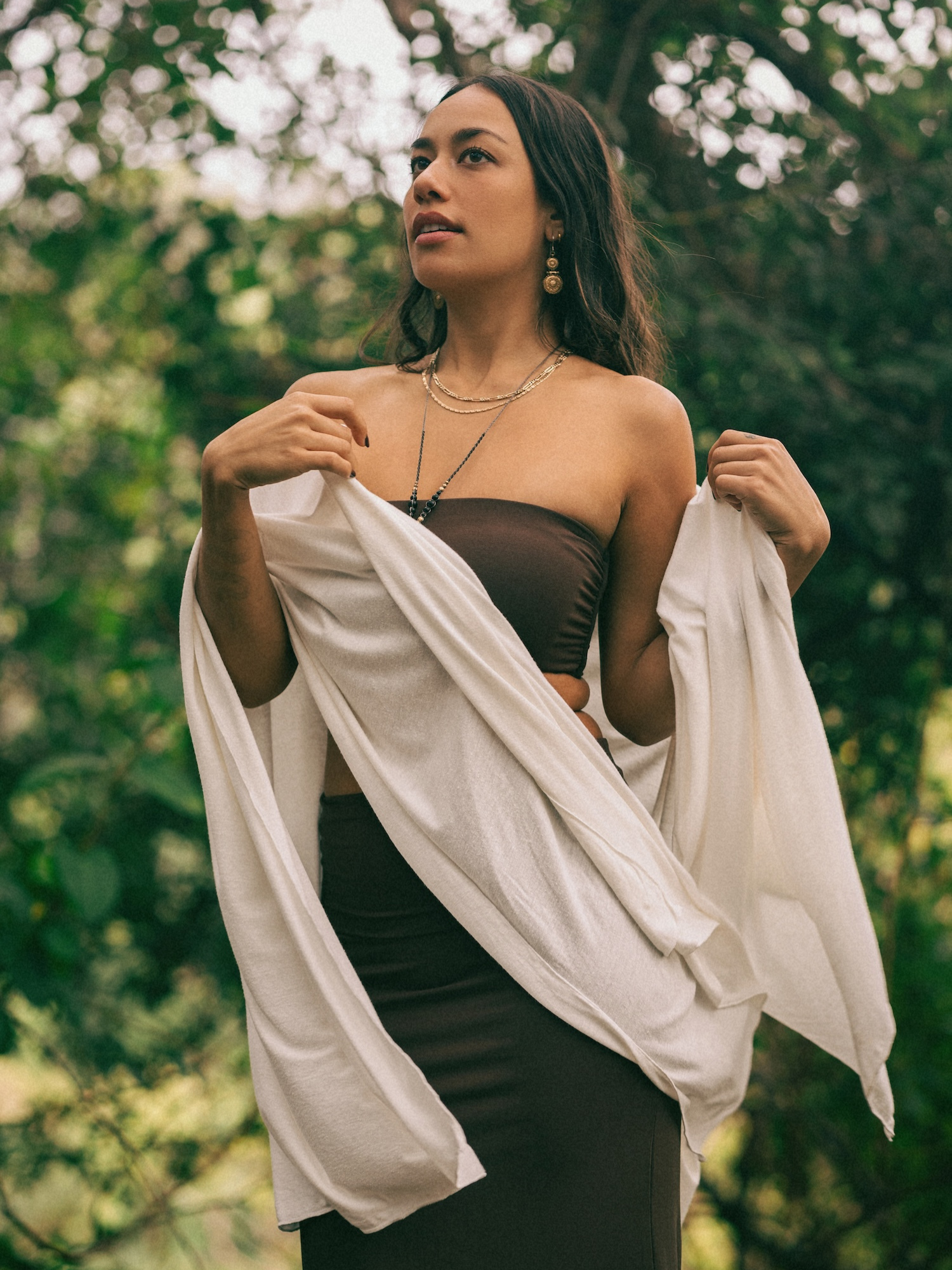 Woman holding a hemp bamboo cream wrap scarf over a brown maxi skirt outdoors with greenery in the background