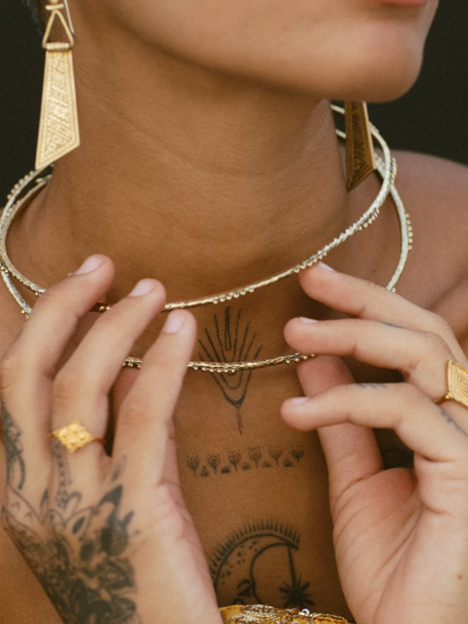 Close-up of a person wearing gold jewelry including earrings, necklace, and rings with tattoos on the neck and hands.