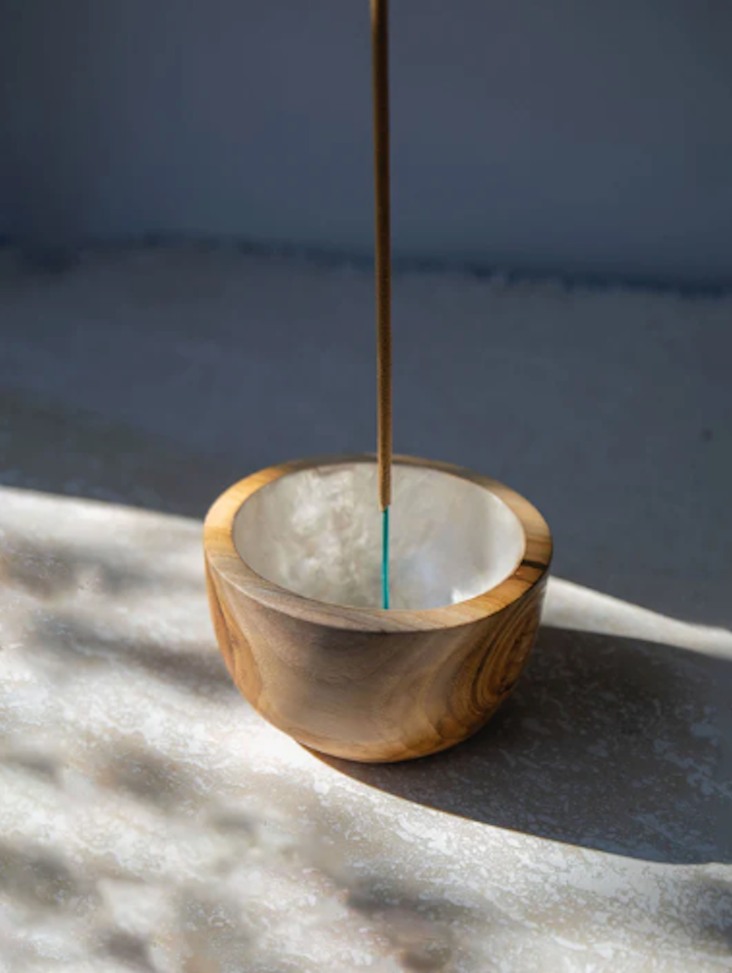 Incense stick in a shell lined wooden cup holder with a white interior on a textured surface.
