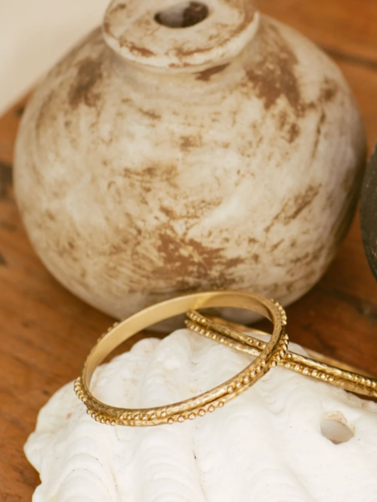 Brass bangles on a white seashell with a textured beige vase in the background